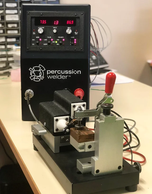 Percussion Welder 858 with its red readouts above the 726 bench fixture and red toggle clamp on a lab bench