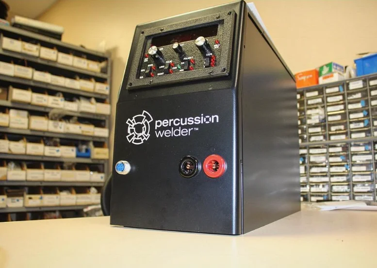 Percussion Welder 858 on a workbench with the red and black weld output jacks and a round connector on its front, parts bins on the shelves behind