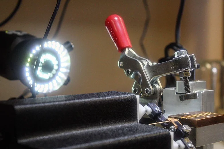 Side view of the bench fixture with a lit ring-light microscope at left, a red-handled toggle clamp and the copper block at right