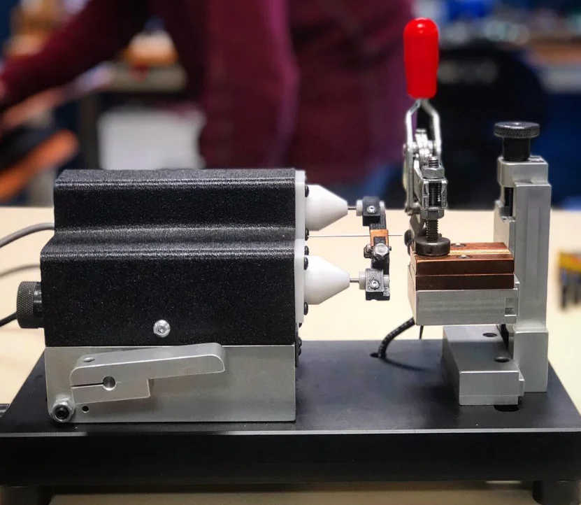 Side view of the 726 bench fixture on a black base: black actuator housings with white cones, an aluminum clamp stand and a red toggle-clamp handle