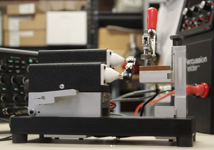 Side view of the 726 bench fixture beside the Percussion Welder, showing the black housings with white cones, the clamp and the red lever