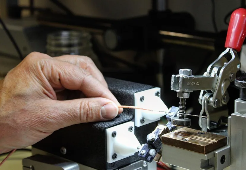 A hand holding a copper wire into the white cone of the bench fixture, with the clamp and copper block at right