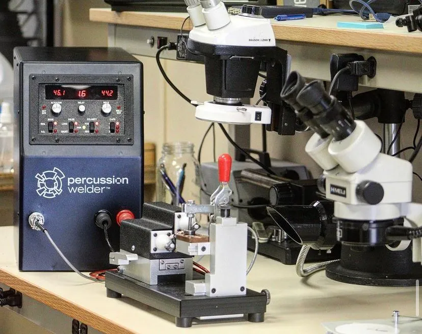 Percussion welder with bench fixture beside two stereo microscopes on a lab bench