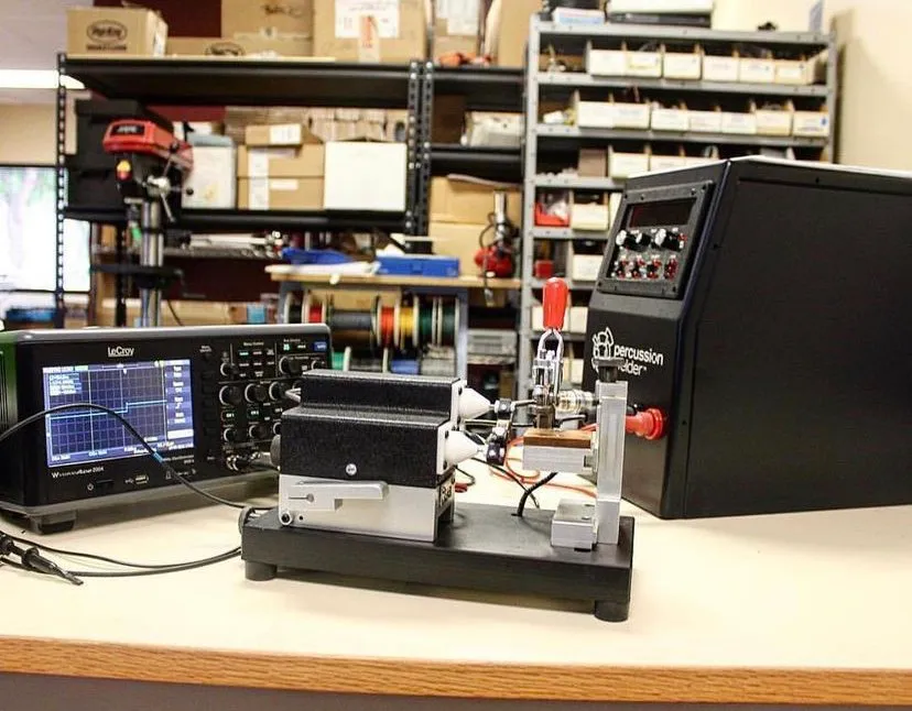 Welder, bench fixture and oscilloscope on a workbench in a shop with shelves of parts