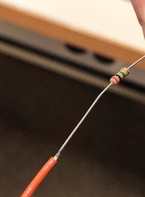 A resistor lead joined to the stripped end of an orange insulated wire, held up in front of a dark background