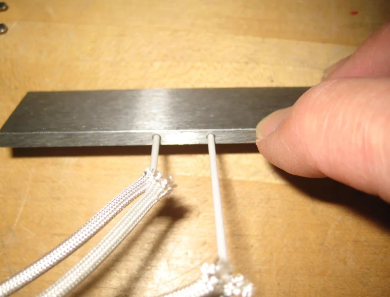 Close-up of two ceramic-insulated thermocouple leads entering the edge of a stainless steel plate, with braided cable and a hand holding the plate