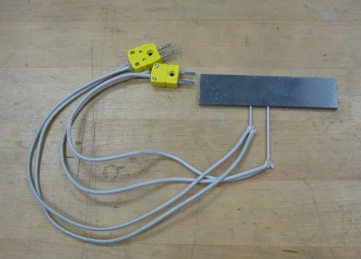 A stainless steel plate with two ceramic-insulated thermocouple leads in its edge, braided insulated cables and two yellow miniature connectors, on a wooden bench