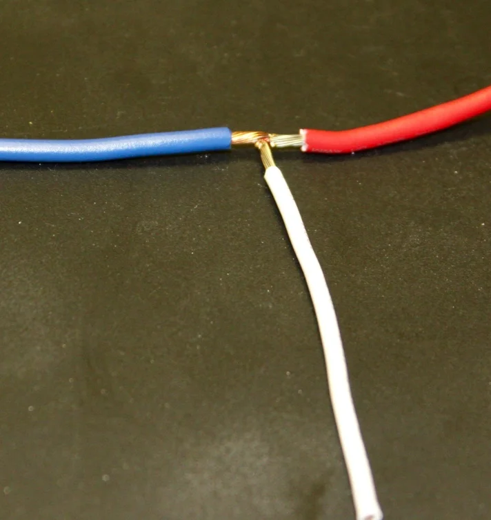 A blue wire and a red wire joined end to end with a white wire welded in at the same junction, on a dark surface